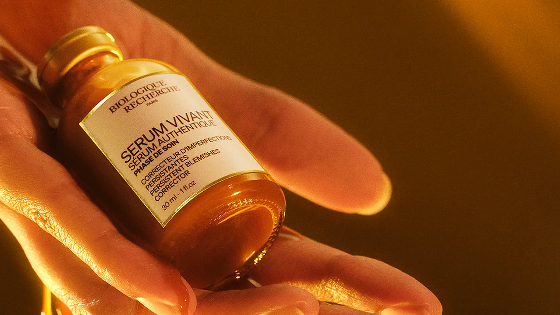 Hand hält den Flakon Serum Vivant von Biologique Recherche, warmes Bernsteinlicht.